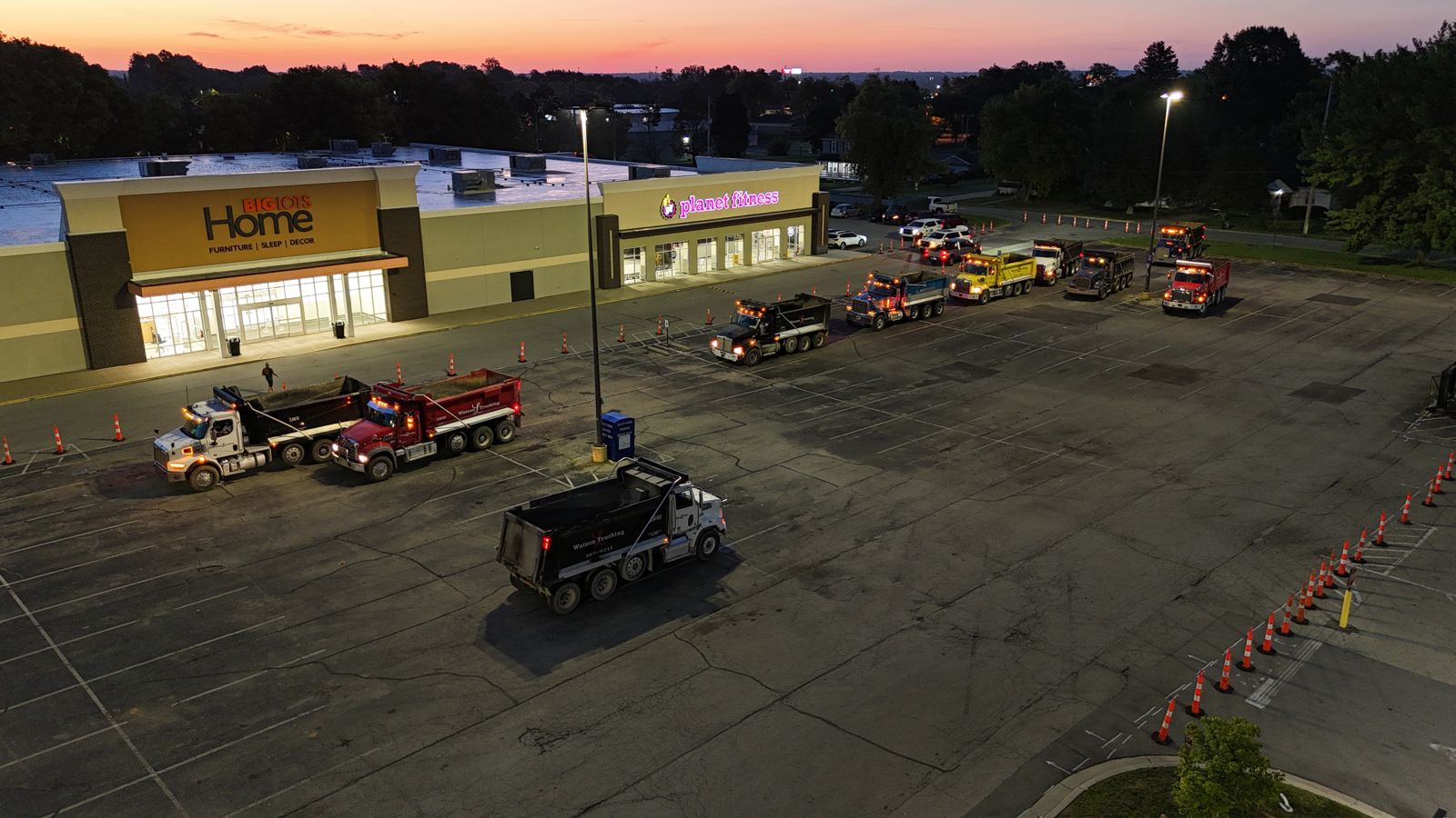 Paving crew and trucks on a big-box store lot at dusk